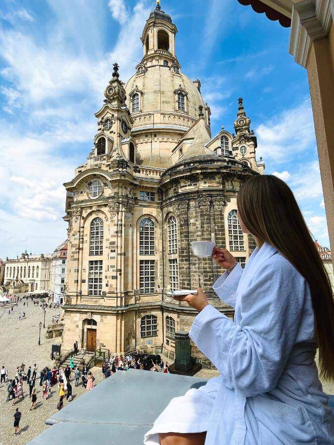 A Woman In A White Robe Holding A Cup And Standing On A Balcony Overlooking A Large Building With Dresden Frauenkirche In The Background